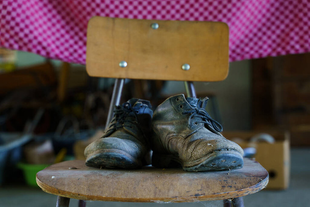 Ένα ζευγάρι παλιά μαύρα δερμάτινα βρώμικα σκονισμένα σκισμένα παπούτσια προειδοποίησης στην καρέκλα στο μπροστινό μέρος της ημέρας σκισμένα πεταμένα μπότες - Φωτογραφία, εικόνα