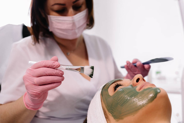 タラソフェイスマスクを適用する美容師。美容師は、スパの治療センターで美しい女性に小さな容器から顔マスクを適用します - 写真・画像