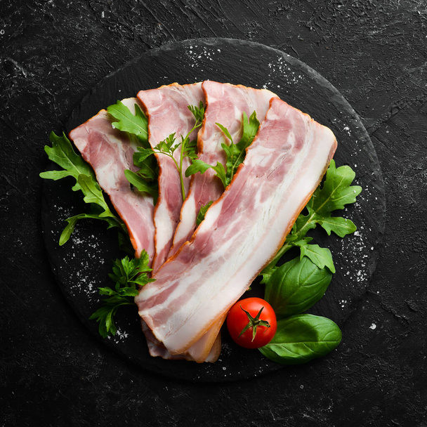 Slices of bacon with arugula and parsley on a black plate. Top view. Free space for your text. - Photo, image