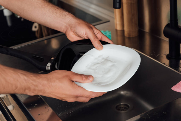 καλλιεργημένη άποψη του ανθρώπου κρατώντας υγρό πιάτο και τηγάνι κοντά νεροχύτη στην κουζίνα  - Φωτογραφία, εικόνα