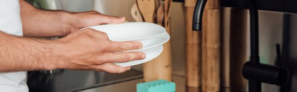 panoramic crop of man holding white plates in kitchen  - Фото, зображення