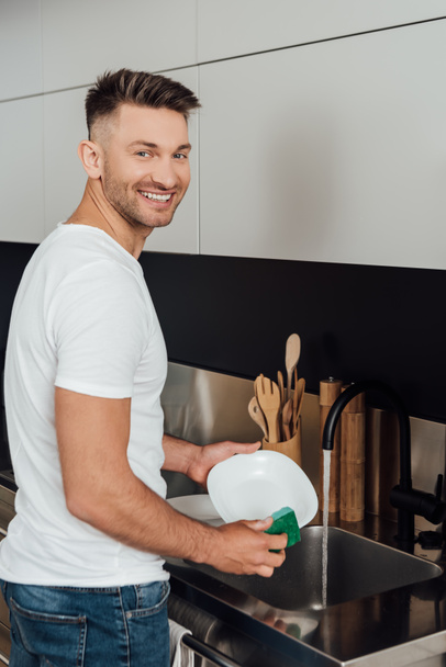 cheerful man smiling while holding sponge and white plate  - Fotoğraf, Görsel
