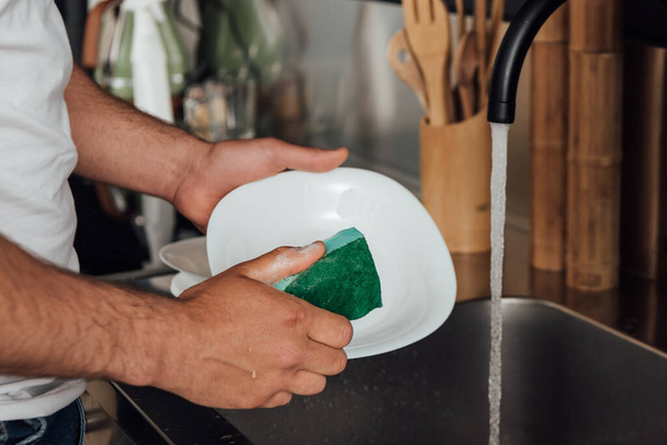 cropped view of man holding sponge while washing white plate  - Foto, imagen