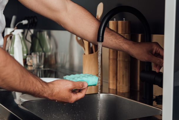 cropped view of man holding sponge near tap water  - Foto, afbeelding
