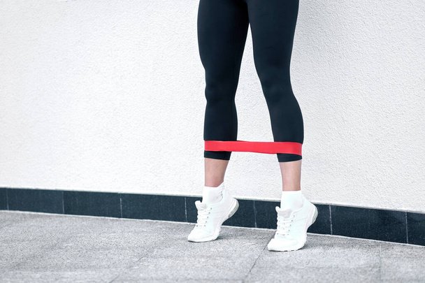 Deportes al aire libre y fitness. Vista de cerca. Chica musculosa fitness con leggins negros y elegantes haciendo ejercicios con caucho rojo fitness cerca de fondo de pared blanco urbano
.                                 - Foto, imagen