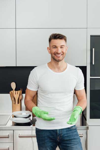 cheerful man in rubber gloves standing near white plates in kitchen   - Foto, Imagen