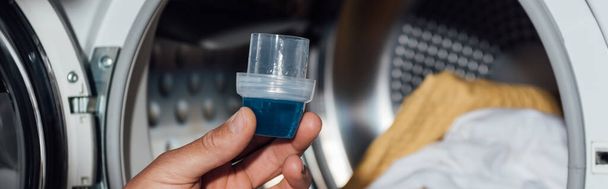 colheita horizontal de homem segurando copo de medição com detergente azul perto da máquina de lavar roupa com roupas sujas
  - Foto, Imagem