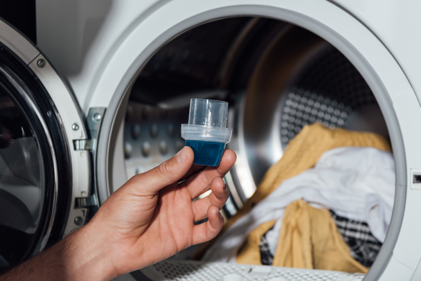 bijgesneden zicht op de man met maatbeker met wasmiddel in de buurt van wasmachine met vuile kleding  - Foto, afbeelding