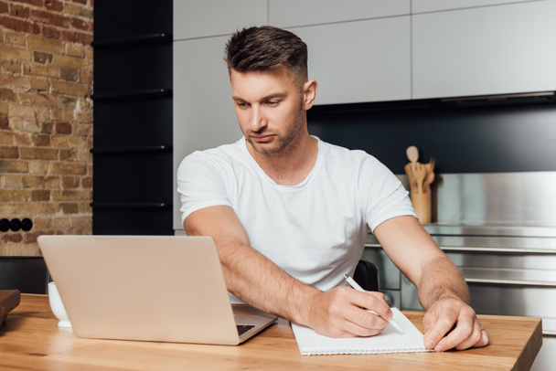συγκεντρωμένος άνθρωπος κρατώντας στυλό και κοιτάζοντας φορητό υπολογιστή, ενώ σε απευθείας σύνδεση μελέτη  - Φωτογραφία, εικόνα
