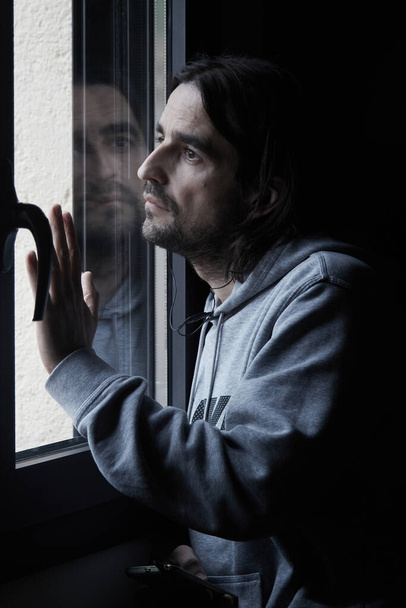 Long-haired man looking out of window sad headphones smartphone - Zdjęcie, obraz