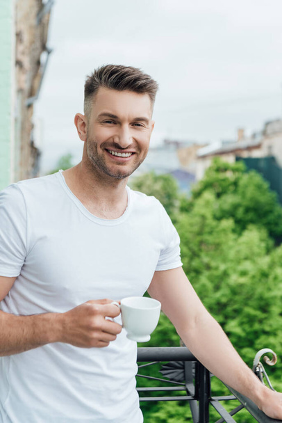 Handsome man smiling at camera while holding cup of coffee on balcony  - Fotó, kép