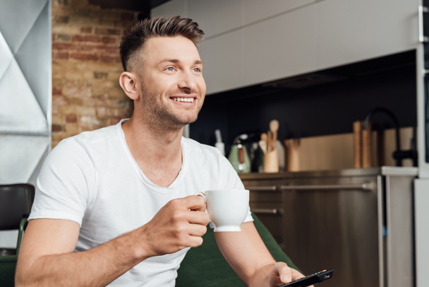 Uśmiechnięty mężczyzna trzyma filiżankę kawy podczas oglądania telewizji na kanapie  - Zdjęcie, obraz