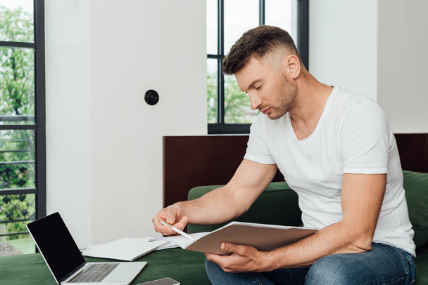 Handsome freelancer holding documents and pen near gadgets on couch at home  - Photo, Image