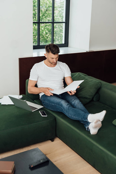 Selective focus of man holding notebook near digital devices and papers on couch  - Foto, Bild