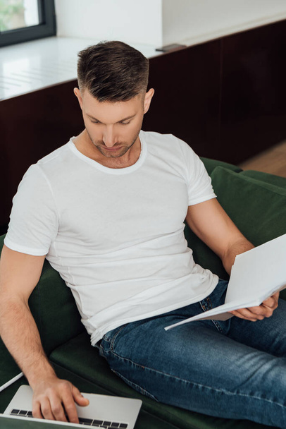 Selective focus of handsome freelancer holding notebook and using laptop at home  - Zdjęcie, obraz