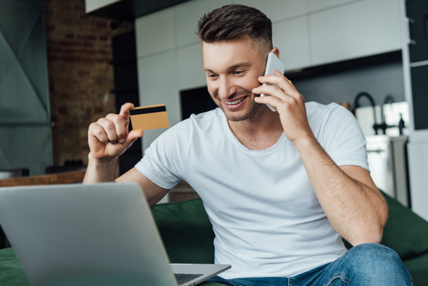 Focus selettivo dell'uomo sorridente che tiene la carta di credito mentre parla su smartphone vicino al laptop in soggiorno
 - Foto, immagini