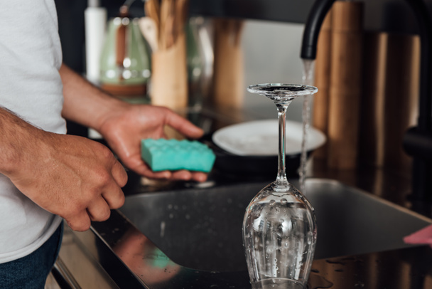 Cropped view of man holding sponge near wet wine glass and kitchen sink  - Foto, Imagem