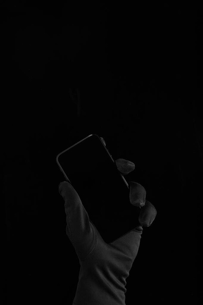 una mano en guante negro sosteniendo un teléfono negro sobre fondo negro
 - Foto, Imagen