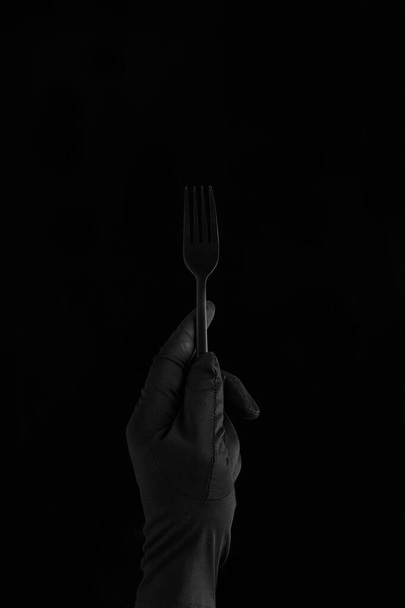 ένα χέρι σε μαύρο γάντι κρατώντας ένα μαύρο πιρούνι σε μαύρο φόντο - Φωτογραφία, εικόνα