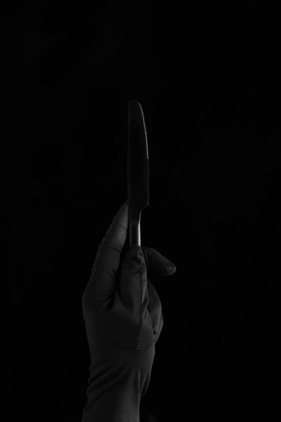 una mano en guante negro sosteniendo un cuchillo negro sobre fondo negro
 - Foto, Imagen