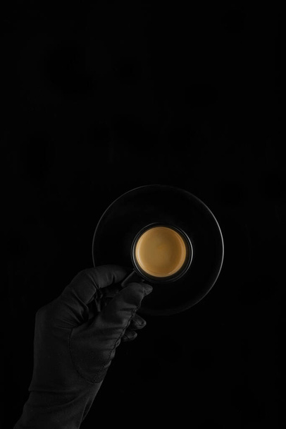 une main en gant noir tenant une tasse de café noire avec expresso sur fond noir
 - Photo, image