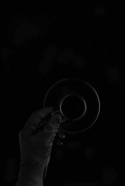 ένα χέρι σε μαύρο γάντι κρατώντας ένα μαύρο άδειο φλιτζάνι καφέ σε μαύρο φόντο - Φωτογραφία, εικόνα