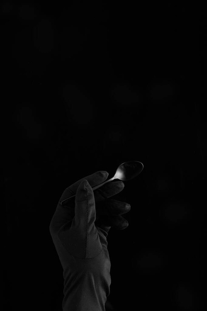 uma mão em luva preta segurando uma colher de chá preta no fundo preto
 - Foto, Imagem
