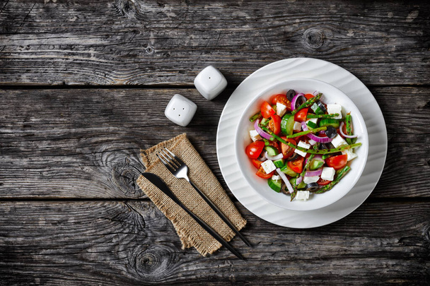 Greek salad with grilled asparagus, cherry tomatoes, cucumbers, black olives, red onion rings with olive oil, feta cheese served on a  plate with cutlery on a wooden table, top view, copy space - Foto, immagini
