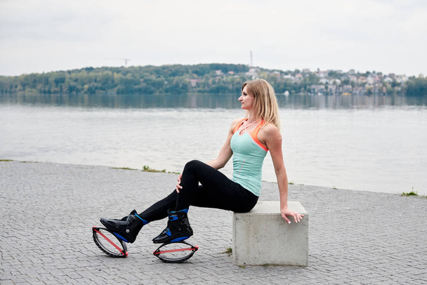 Young blond fit slim woman in kangoo jumps, sitting by city lake in summer. Fitness trainer, wearing black leggings and mint top, doing exercises, posing with her legs up in the air. Healthy lifestyle - Foto, Imagem