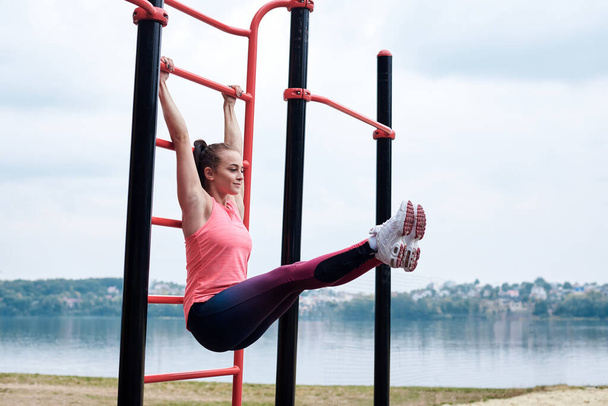 Young brunette woman, wearing pink top and blue leggings, hanging on wall bars outside, lifting legs up by city lake. Slim sportswoman, doing stretching fitness exercises. Healthy lifestyle concept. - 写真・画像
