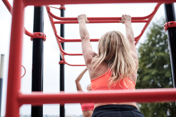 Young women, wearing pink tops and dark leggings, hanging on horizontal bars outside by city lake. Slim sportswomen, doing fitness exercises. Healthy lifestyle concept. Strength training. - Foto, Imagem