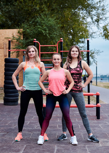 A group of three young women, wearing colorful sports outfit,doing fitness exercises outside in summer.Workout power female training to loose weight and body shape at nature. Healthy lifestyle concept - Фото, изображение