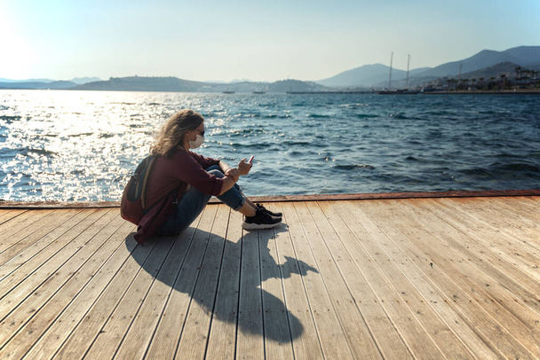 Young girl woman in a mask and sunglasses sits on a wooden pier on the seashore with a smartphone in her hands. Coronavirus pandemic, weakening of protective measures. - Photo, image