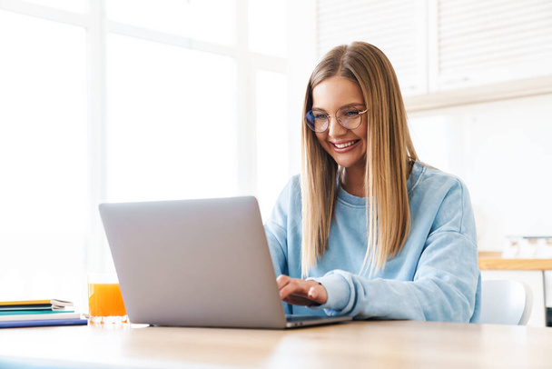 Image of cheerful charming woman in eyeglasses smiling while working with laptop at home - Zdjęcie, obraz