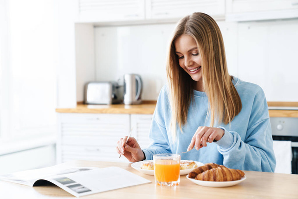 Image of cheerful young woman with blonde hair smiling while having breakfast at modern kitchen - Zdjęcie, obraz