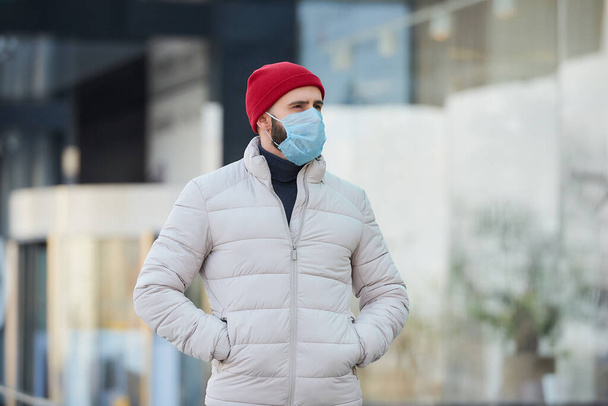 Mężczyzna rasy kaukaskiej noszący medyczną maskę na twarz, aby uniknąć rozprzestrzeniania się koronawirusu (COVID-19). Facet w masce chirurgicznej na twarzy z powodu pandemii na ulicy.. - Zdjęcie, obraz