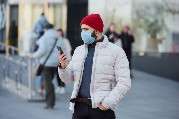 拡散性コロナウイルス(COVID-19)を避けるために医療面マスクを着用している白人男性。町の中心部にスマートフォンを持っている顔に外科用マスクをした男. - 写真・画像