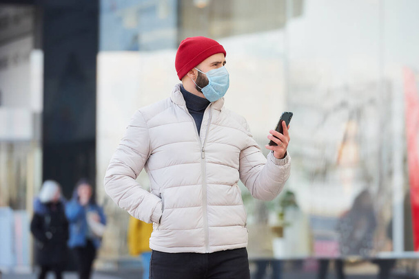 A man wearing a medical face mask to avoid the spread coronavirus (COVID-19). A guy with a surgical mask on the face because of the pandemic looking around holds a smartphone in the center of the city - Photo, Image