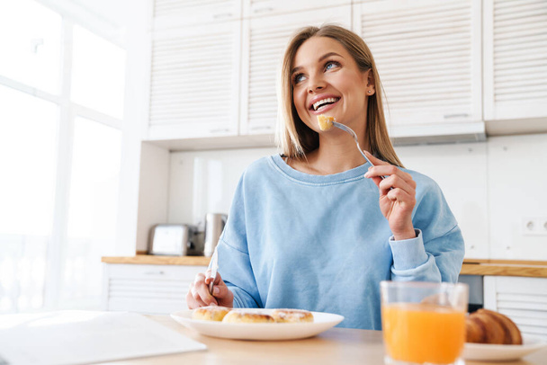 Image of cheerful young woman with blonde hair smiling while having breakfast at modern kitchen - Φωτογραφία, εικόνα