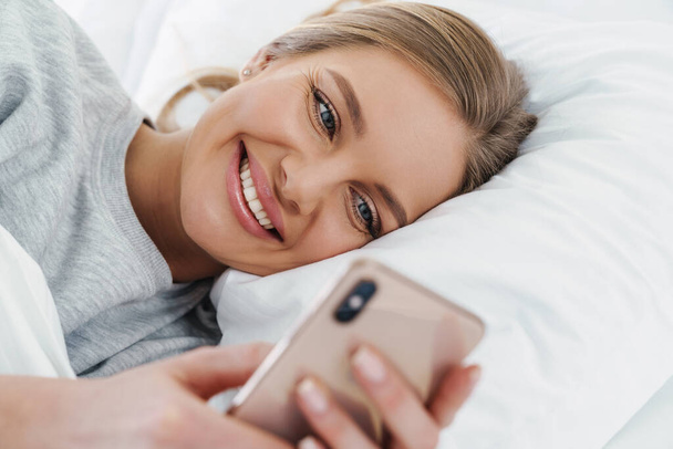Image closeup of cute joyful woman smiling and using cellphone while lying after sleep in bed - Fotó, kép