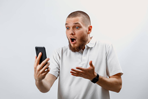 Πορτρέτο ενός θετικού όμορφου γενειοφόρου άνδρα με λευκό μπλουζάκι που μιλάει σε βιντεοκλήση χρησιμοποιώντας ένα smartphone. απομονωμένη. συναισθήματα - Φωτογραφία, εικόνα
