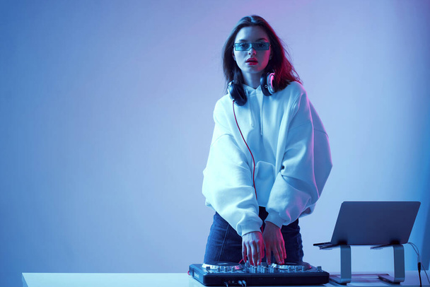 Cool young girl DJ mixes music on a mixing console and laptop, in stylish clothes, glasses on a neon background. - Foto, imagen