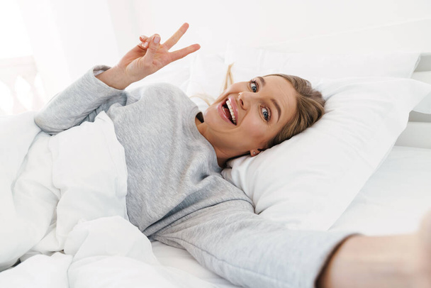 Obraz uroczej uśmiechniętej kobiety gestykulującej znak pokoju i robiącej zdjęcie selfie leżącej po śnie w łóżku - Zdjęcie, obraz