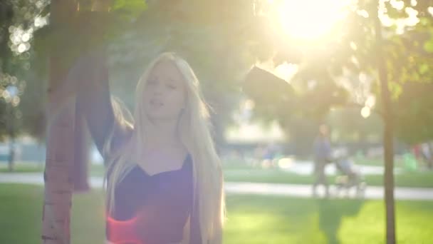 Young Caucasian girl with long blond hair in sun in city park touches green leaves with her hand. Teenage model in stylish casual black blouse turns around, smiles sweetly, poses. Fashion and ecology. - Video