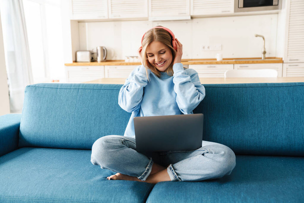 Imagen de una linda mujer sonriente usando un portátil y auriculares inalámbricos mientras está sentada en el sofá en casa
 - Foto, imagen