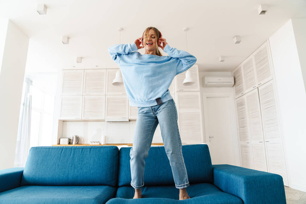 Image of cute smiling woman dancing on couch while using wireless headphones at home - Zdjęcie, obraz