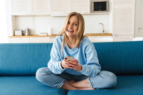 Image of young cheerful woman using smartphone and laughing while sitting on sofa at home - Foto, afbeelding