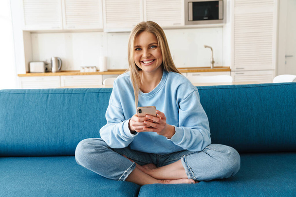 Image of young cheerful woman using smartphone and smiling while sitting on sofa at home - Foto, Imagen