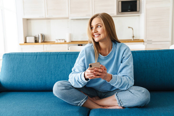 Image of young cheerful woman using smartphone and smiling while sitting on sofa at home - Foto, afbeelding