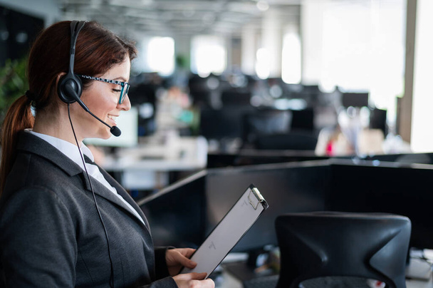 Friendly business woman in a suit talking to a customer on a headset. Female call center employee answers customer questions according to instructions. Support service. - Foto, Imagen